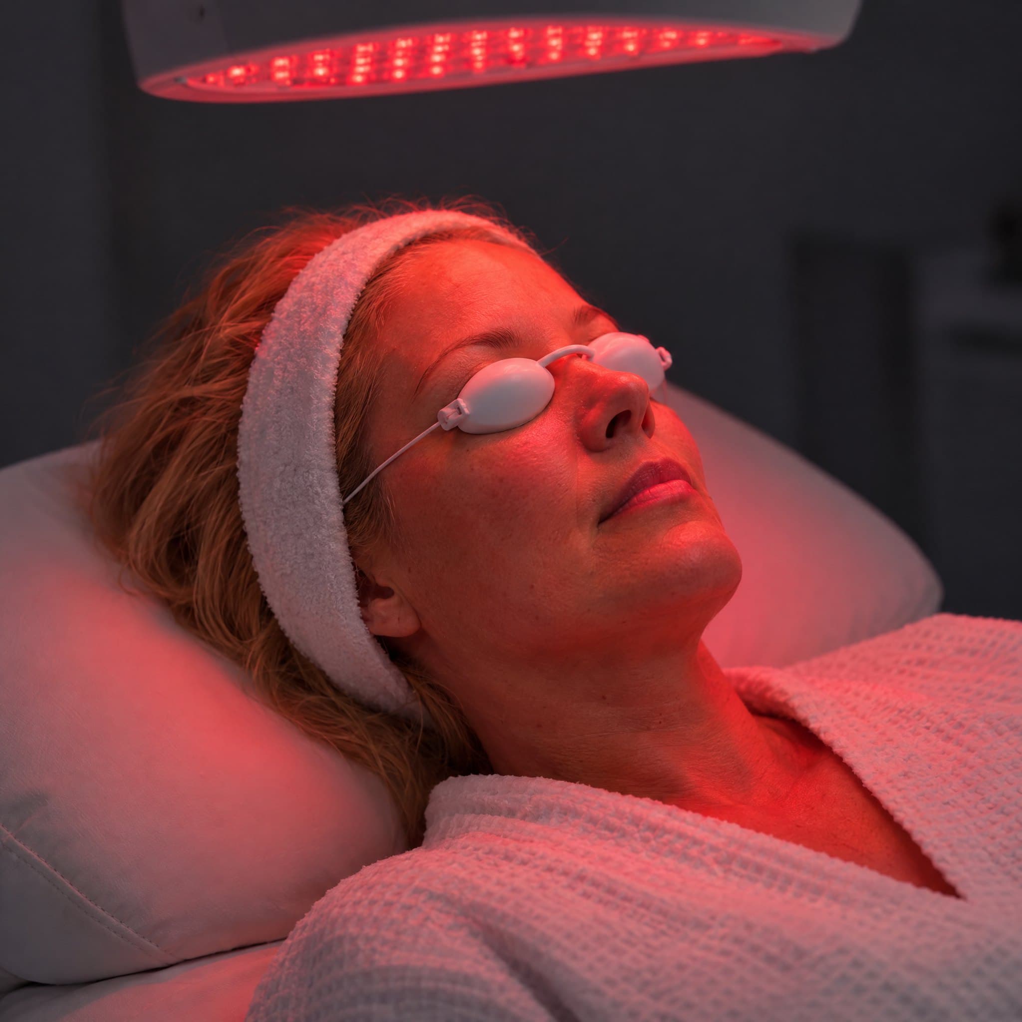 A client under a red LED light therapy panel wearing protective eye shields