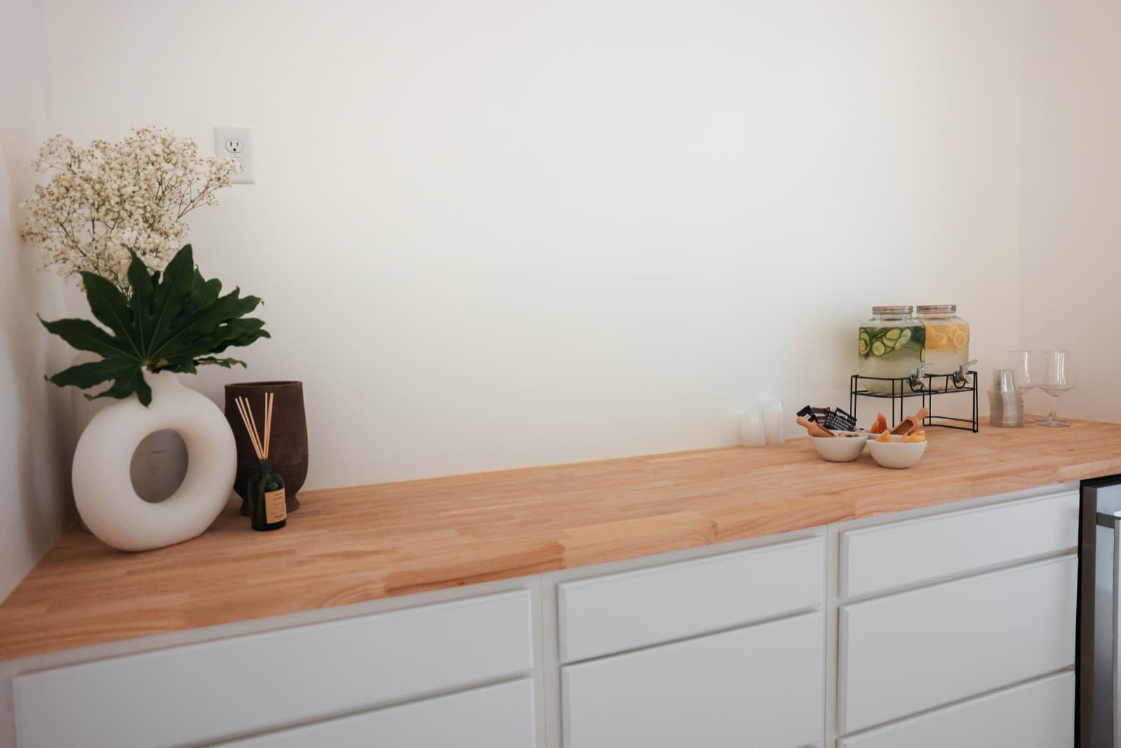 Ora's refreshments counter — infused water, a vase of flowers and a reed diffuser on a wood countertop