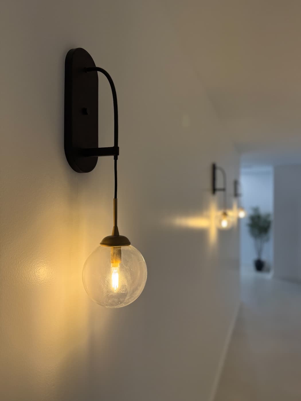 A glass globe sconce glowing warm on a hallway wall at Ora, more lights trailing down the corridor behind it