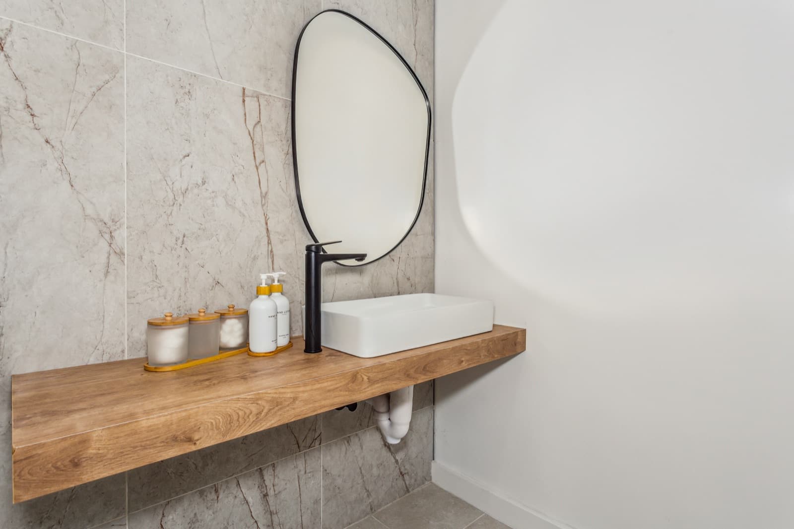 A washroom vanity at Ora — a wood shelf, vessel sink and pebble-shaped mirror against marble tile